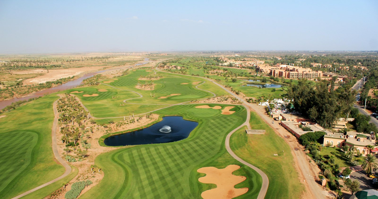 Vue aérienne de l'extension du parcours de la Palmeraie à Marrakech : fairways verts, plan d'eau central, bunkers en forme libre et palmeraie en arrière-plan.