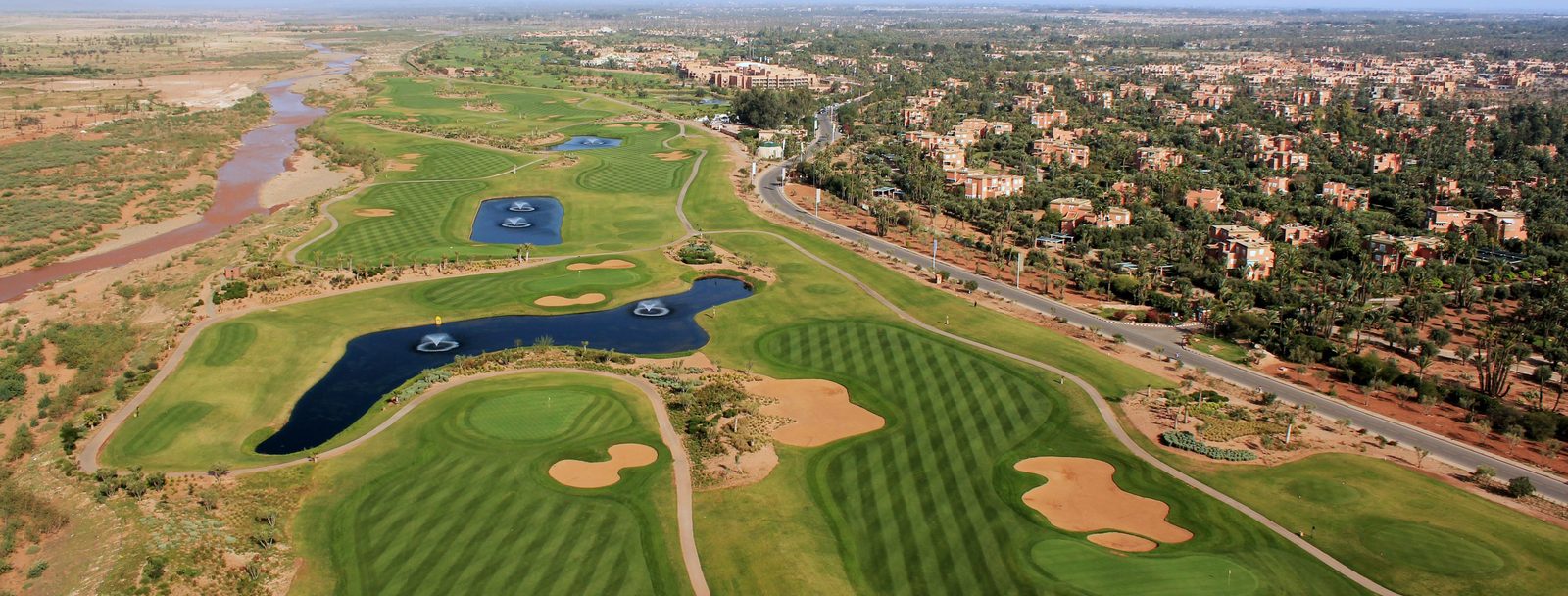Panoramique de l'extension de la Palmeraie : la succession des fairways, des plans d'eau et de l'oued, avec Marrakech au loin.